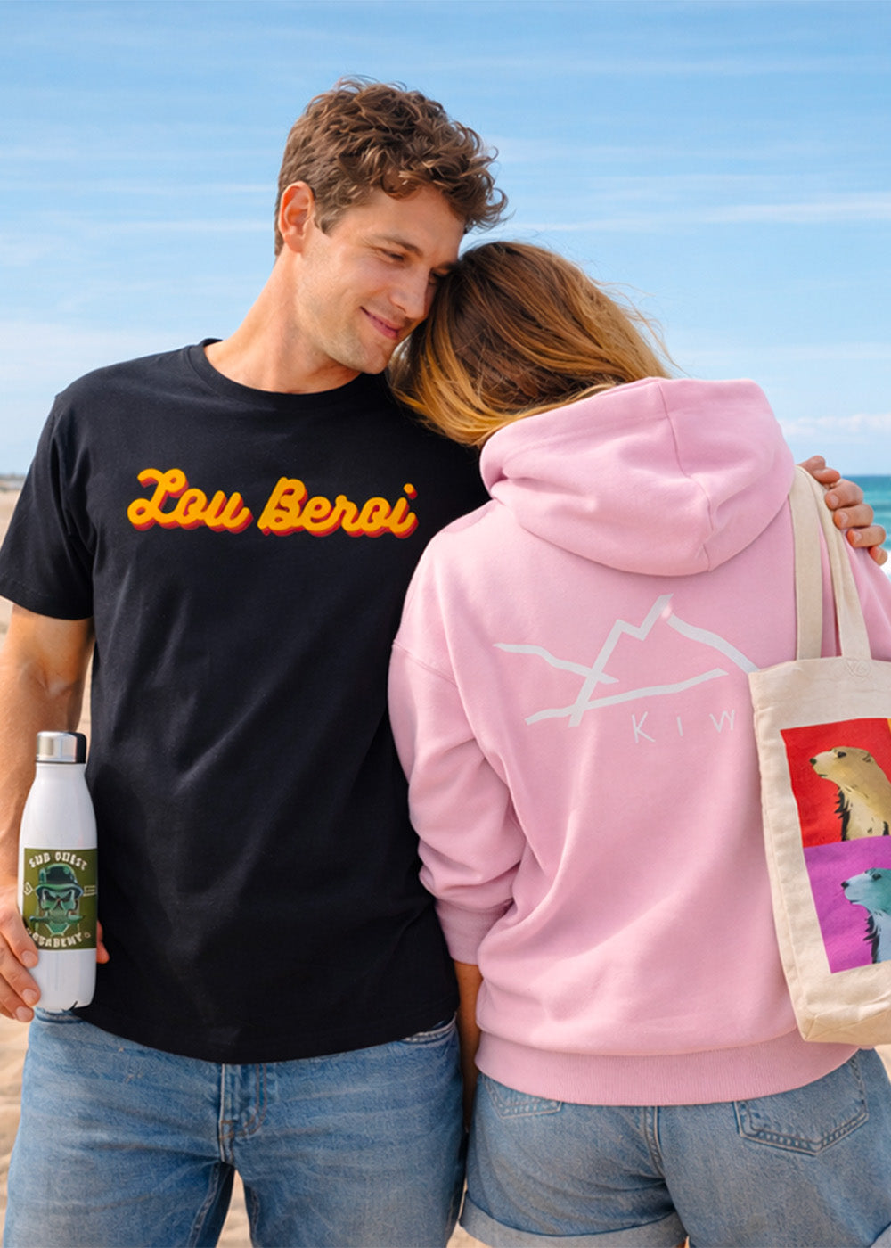 Couple portant un t-shirt noir Lou Beroi et un sweat rose Kiwa sur la plage d’Hossegor avec tote bag et gourde