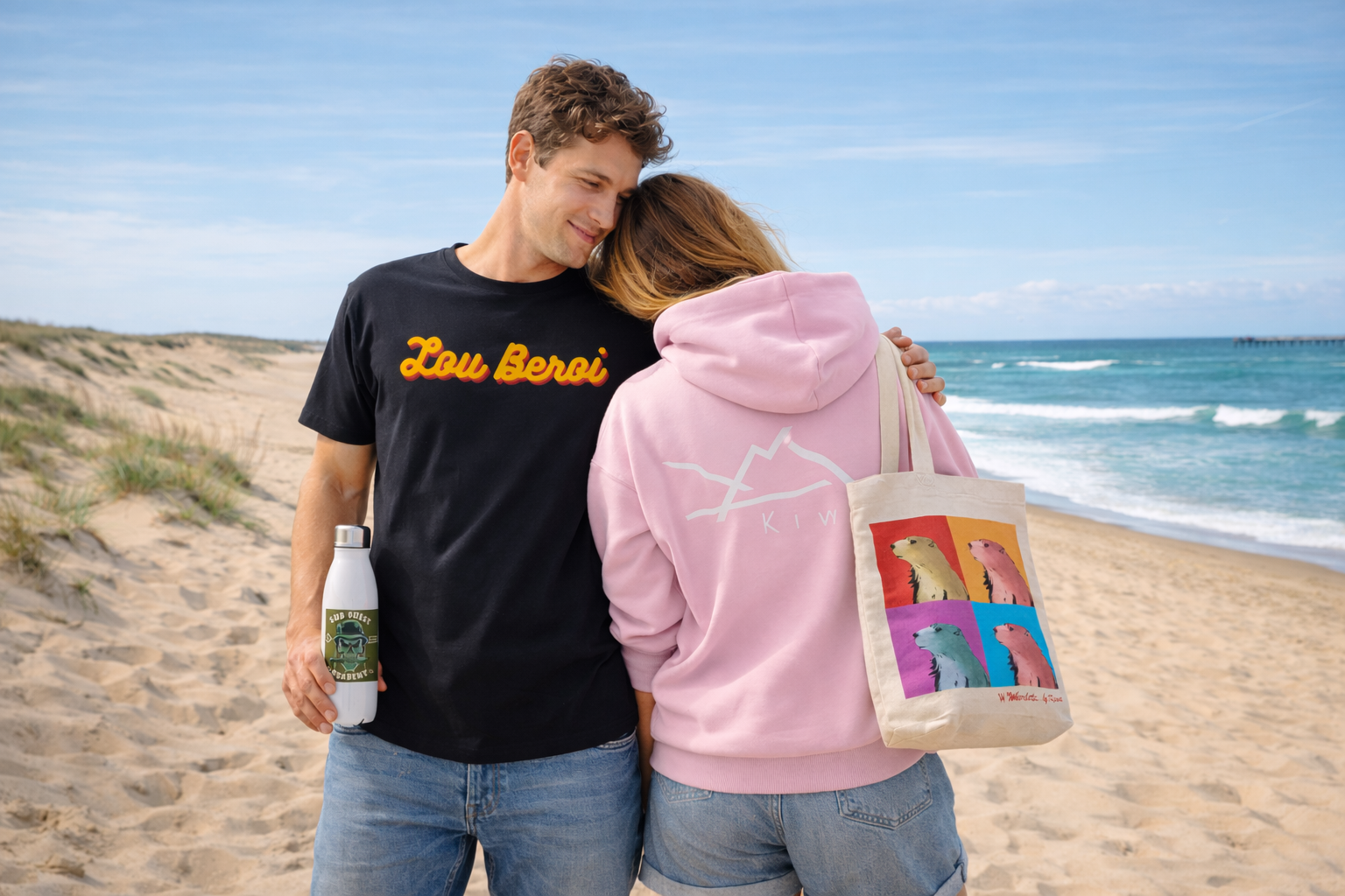 Couple portant un t-shirt noir Lou Beroi et un sweat rose Kiwa sur la plage d’Hossegor avec tote bag et gourde